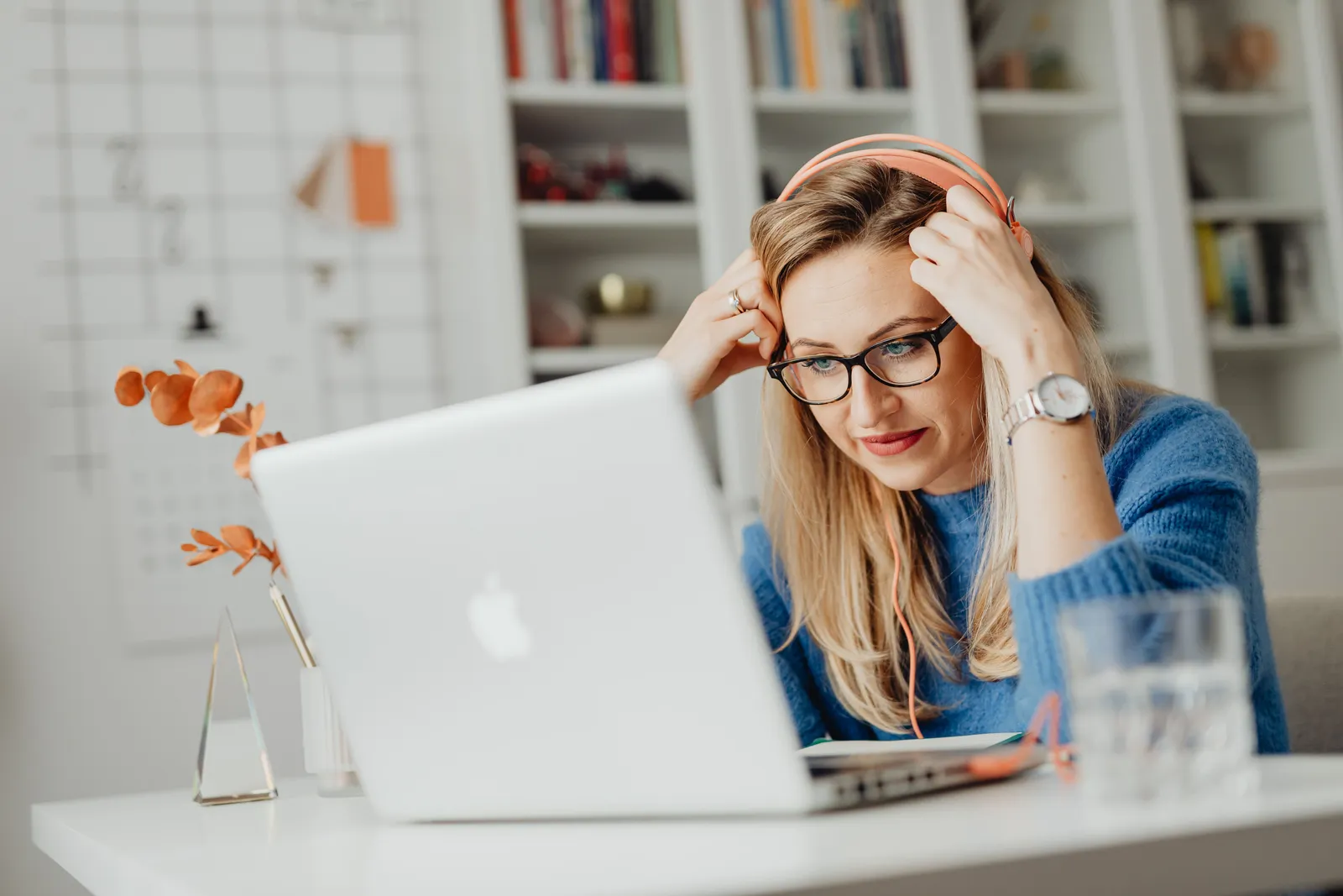 Blond woman wearing eyeglasses and headphones, working on a laptop indoors at a desk
