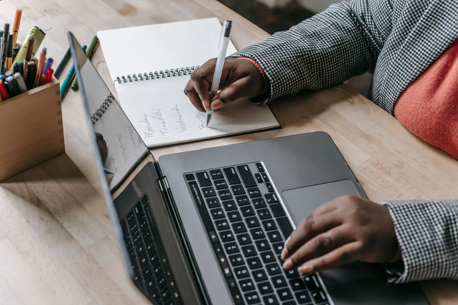 Crop unrecognizable female in formal wear writing notes in notepad while working on laptop