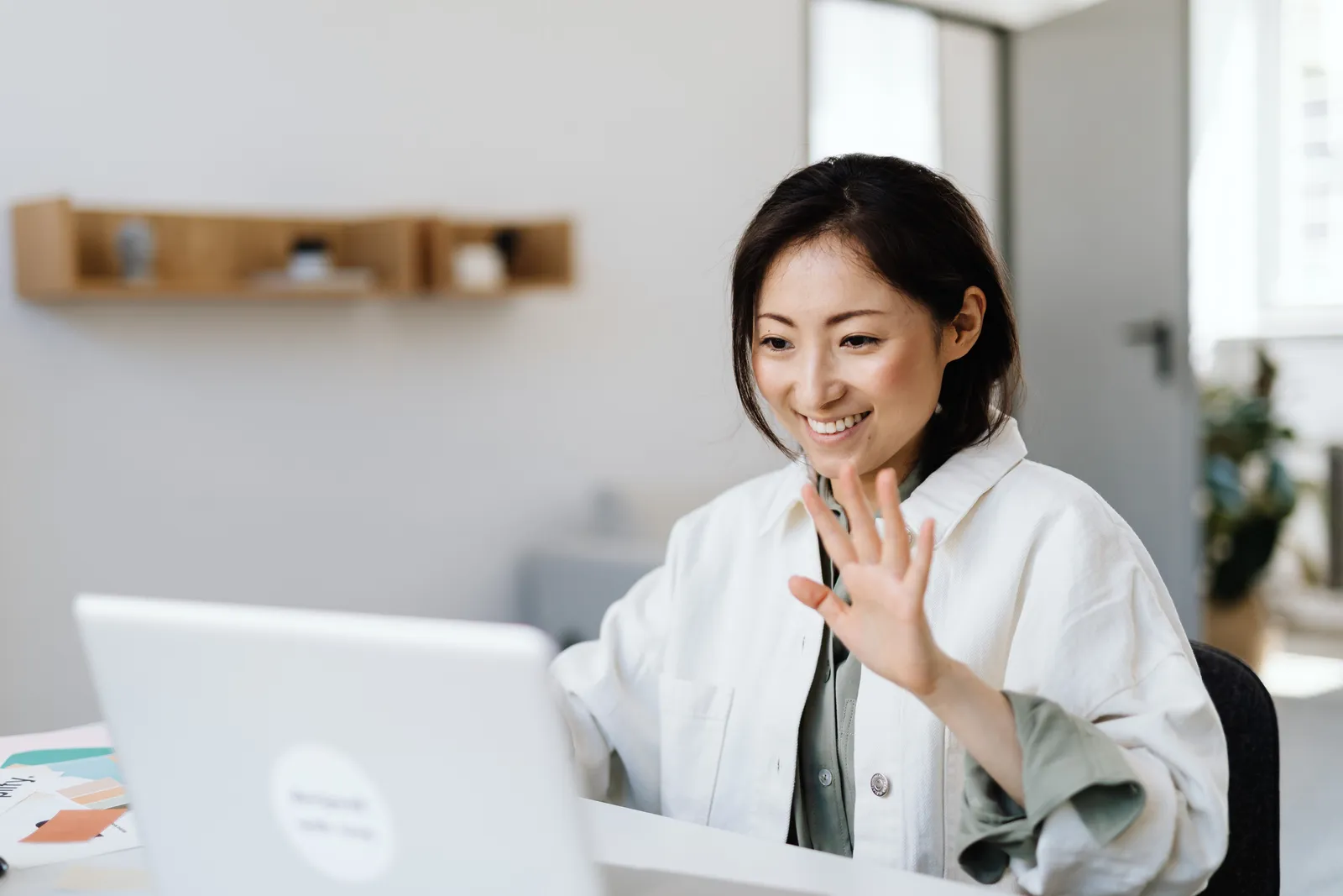 Smiling woman waves at laptop during a video call in a modern office