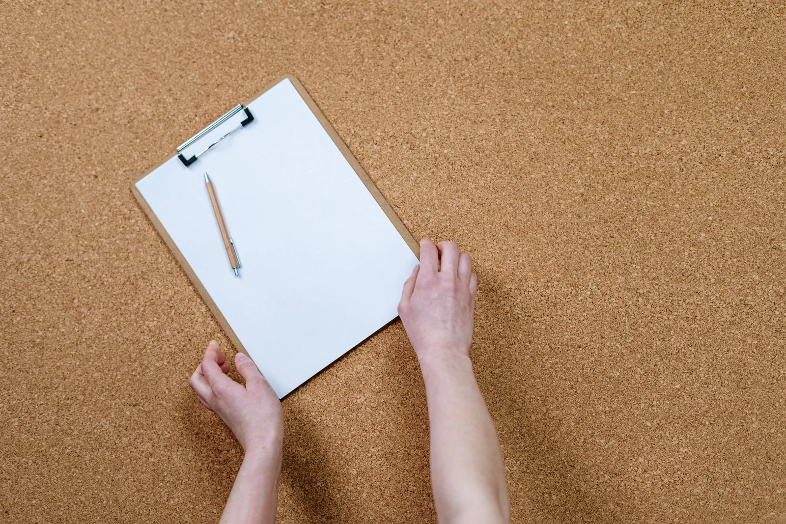Hands holding a clipboard with paper and pencil on a cork board. Ideal for office or school themes