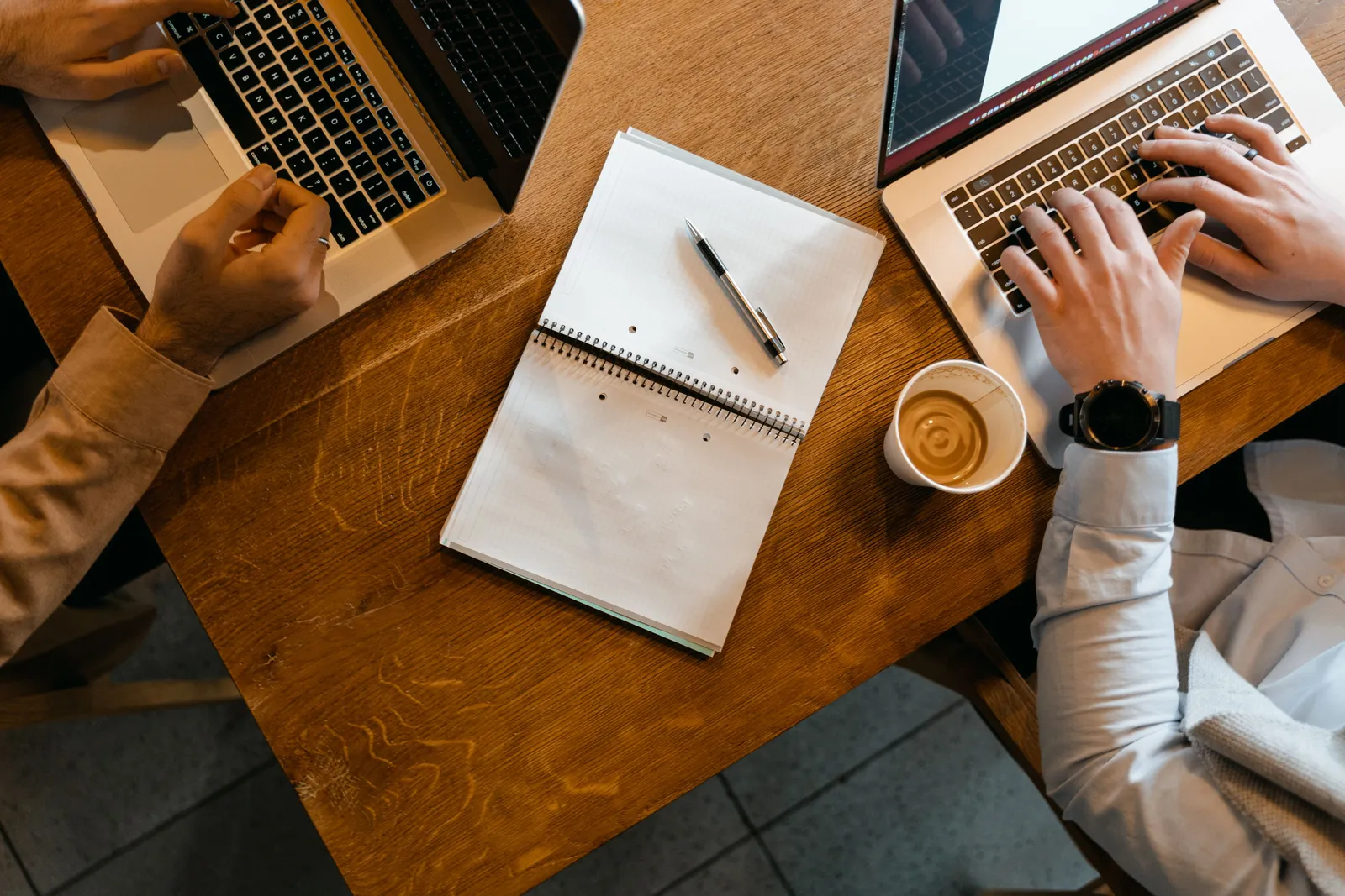 Overhead shot of coworkers using laptops with notes and coffee at a wooden desk