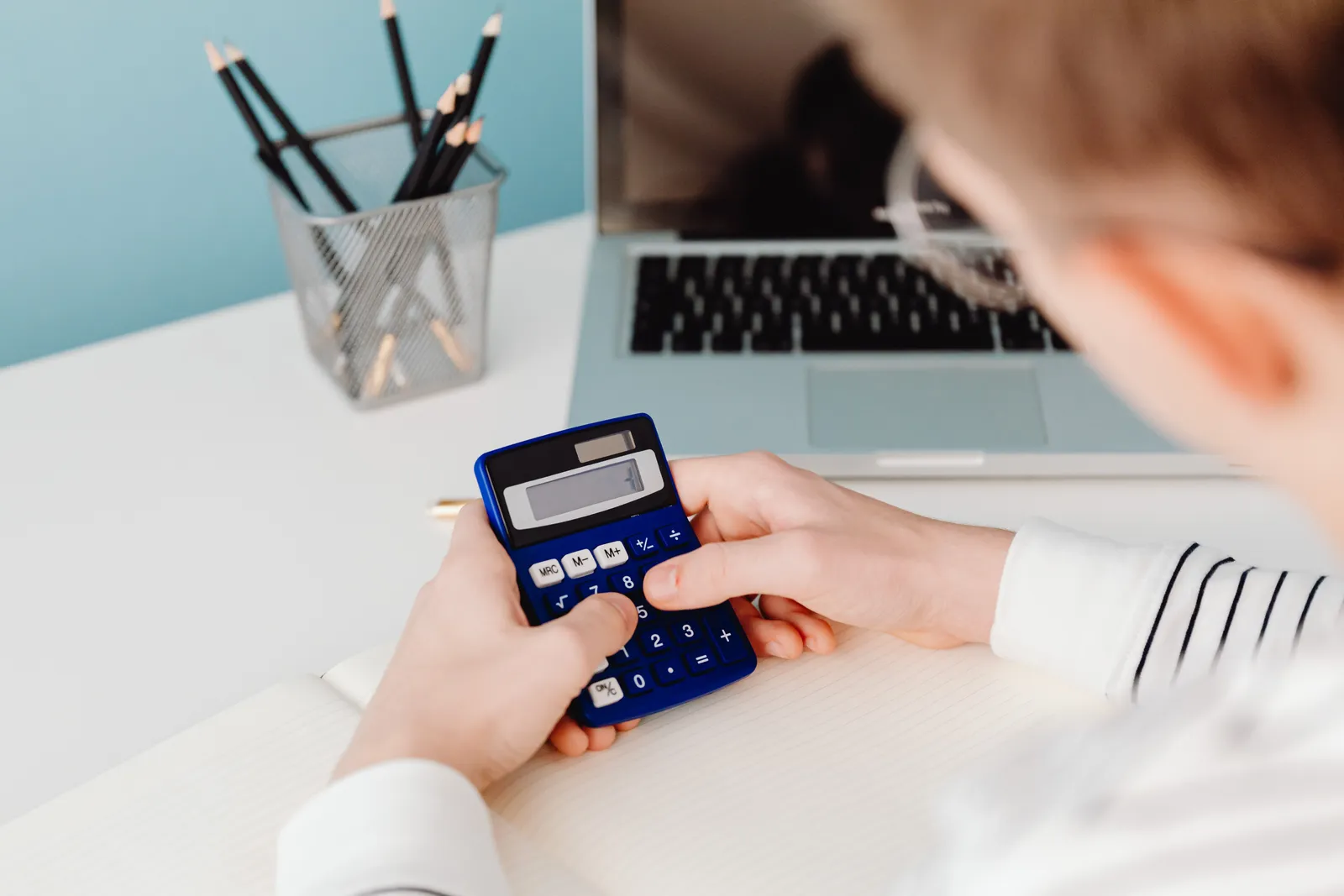 Close-up of hands using a blue calculator at a desk with a laptop and stationery