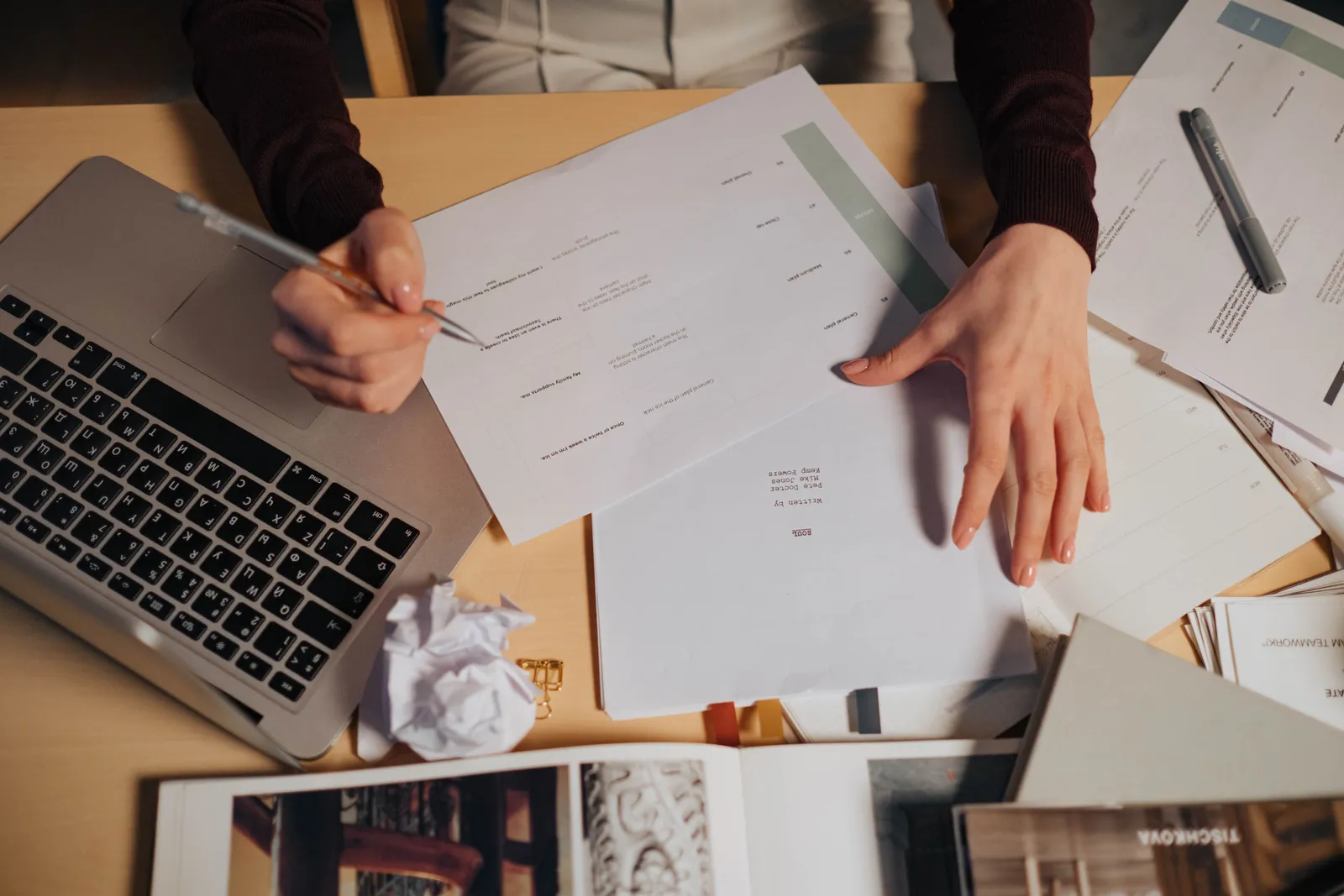 A person taking notes and working on a laptop surrounded by documents on a desk