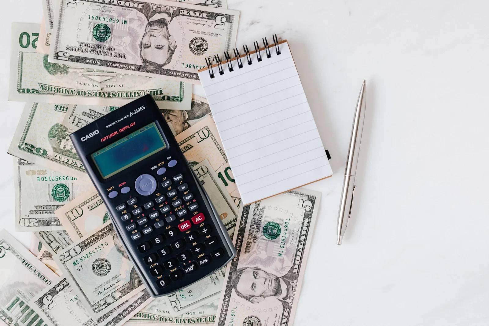Top view of electronic calculator and notepad placed over United States dollar cash money together with metallic pen for