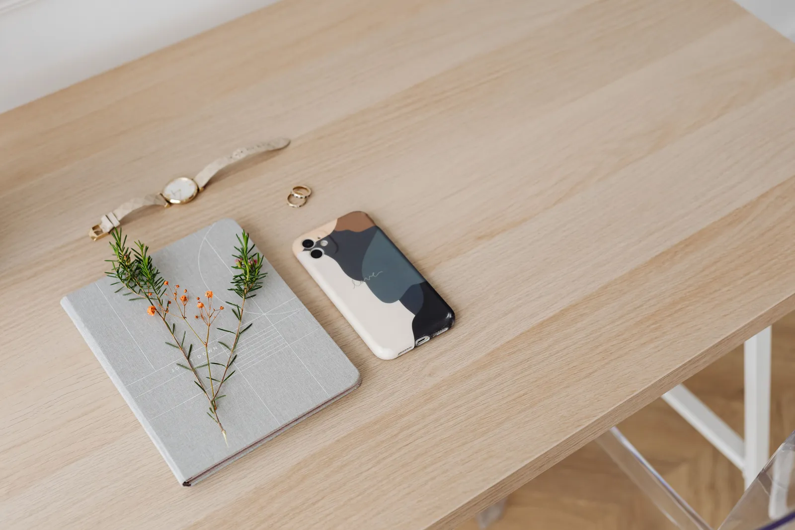 A minimalist desk with a phone, notebook, watch, ring, and plant sprig, showcasing elegance and simplicity