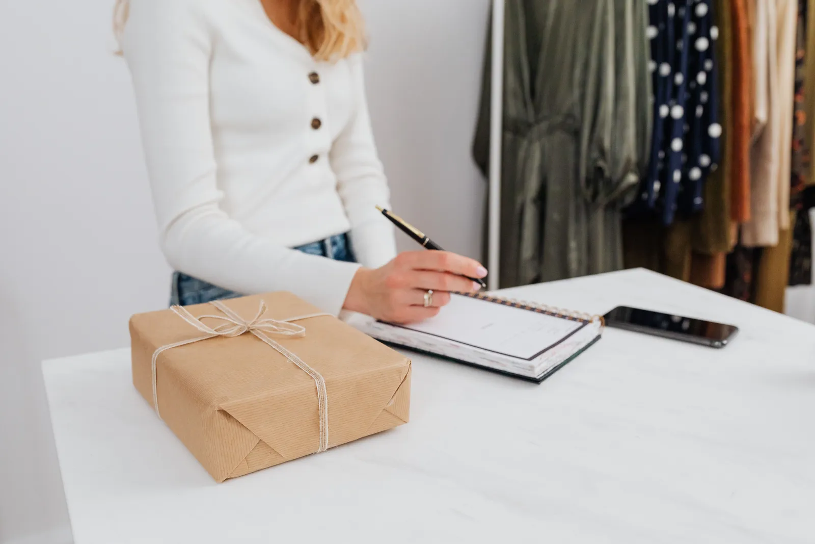 Woman writing in notebook beside a wrapped gift box on a white table indoors