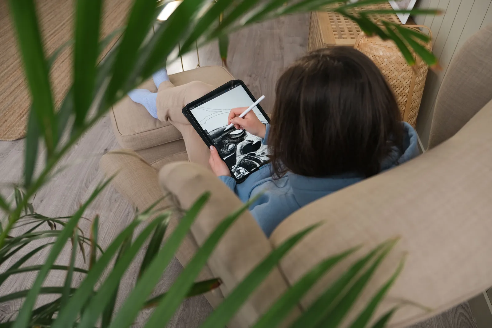 A person drawing on a digital tablet while lounging on a sofa, surrounded by plant leaves