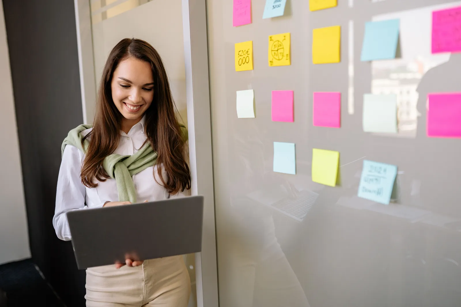 Smiling woman uses laptop in office, planning with colorful sticky notes on glass wall