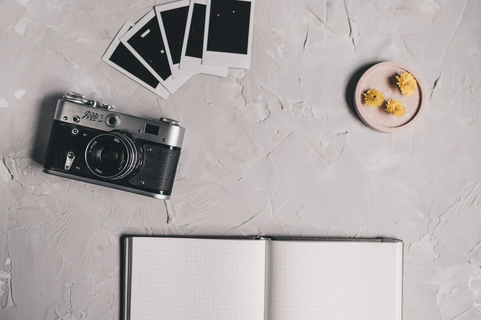 A vintage camera and polaroids on a textured desk with an open notebook and flowers