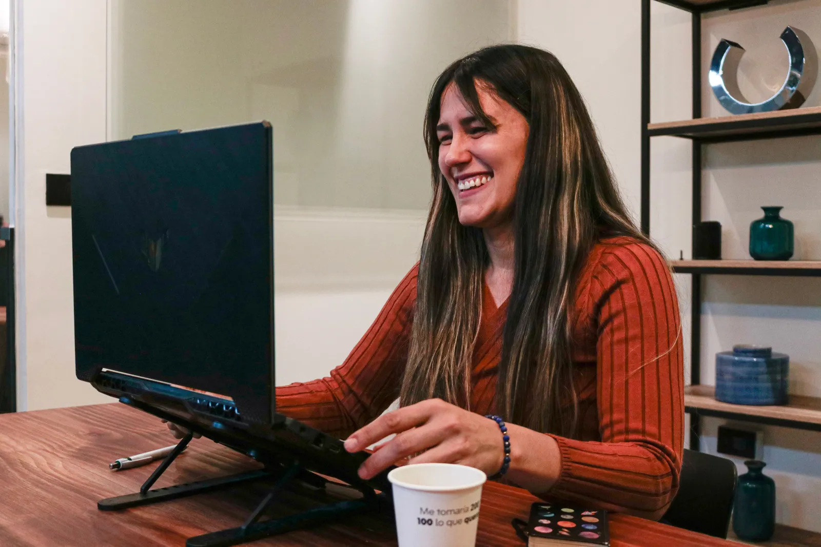 Smiling woman in red sweater enjoying a video call at home office setup with laptop and coffee