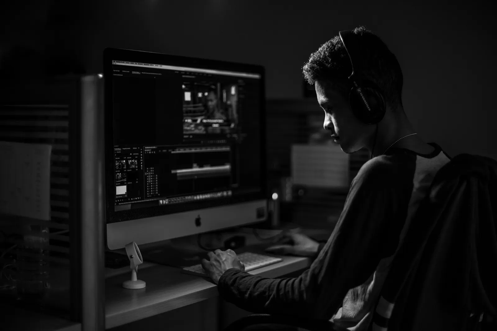 A man wearing headphones working on video editing on a computer in a dimly lit office