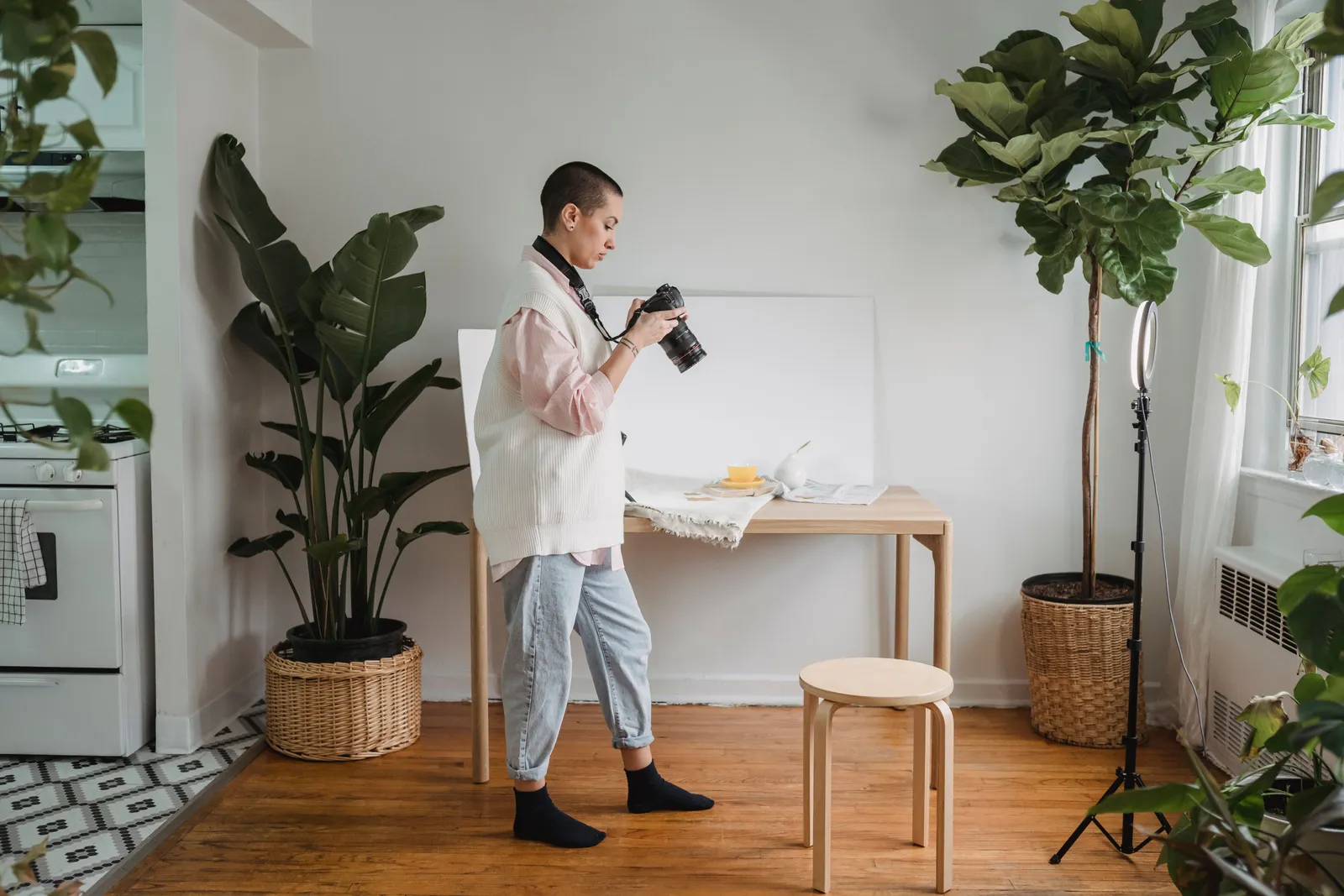 Side view of young woman watching digital photo camera on floor between potted plants in light house