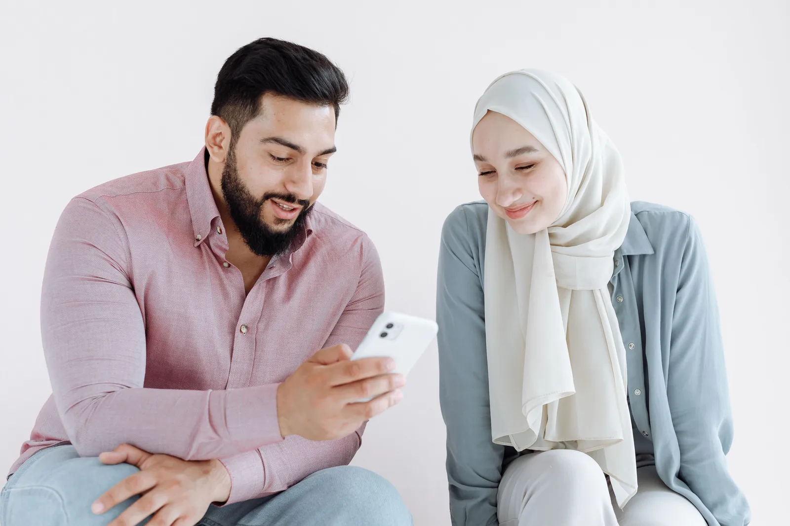 A bearded man and a woman in hijab share a mobile phone moment indoors, conveying modern communication