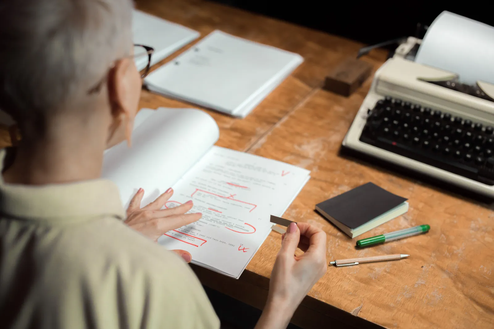 An adult woman reviewing a script with red pen marks at a wooden desk with a typewriter