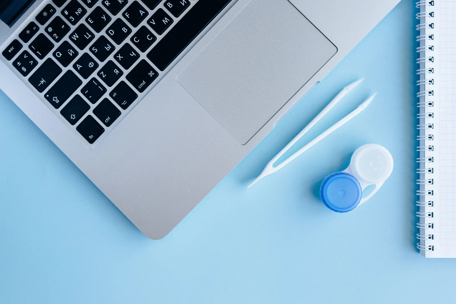Top view of contact lens case, laptop, and notebook on a blue surface