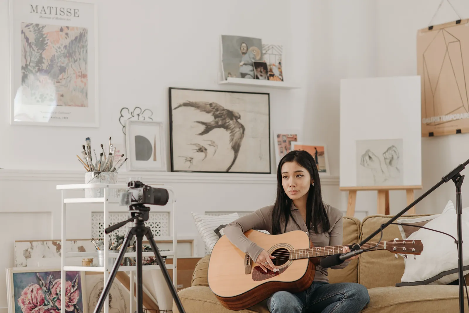 Woman strumming acoustic guitar in a modern living room setup with camera and art pieces
