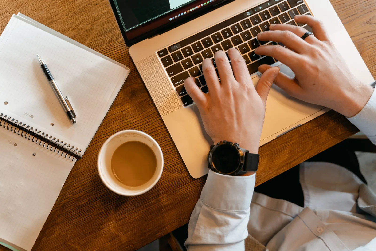 Hands typing on a laptop next to a cup of coffee and a notebook on a wooden table