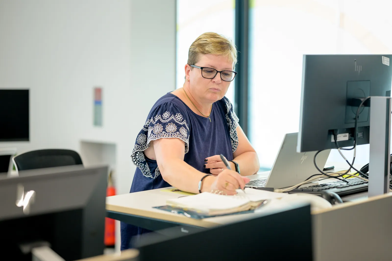 Professional woman wearing eyeglasses working at an office desk with a laptop and notebook