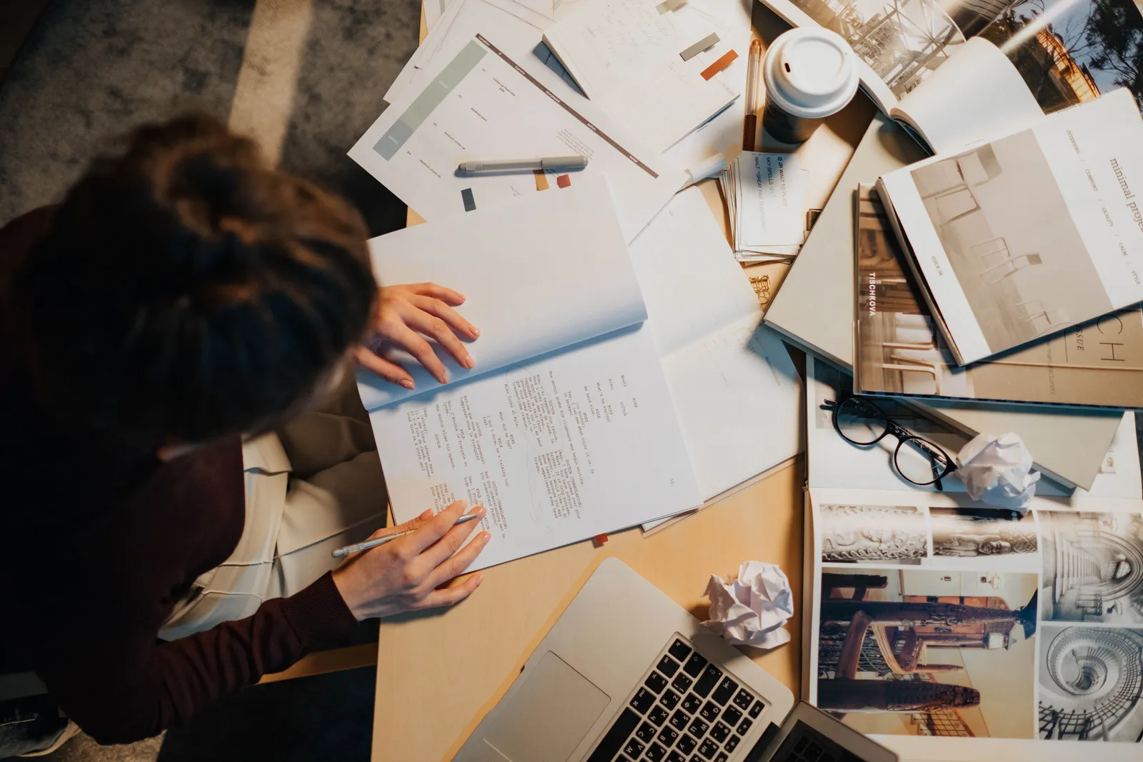 Overhead view of a cluttered work desk with a person writing notes, surrounded by papers, laptop, and coffee cup