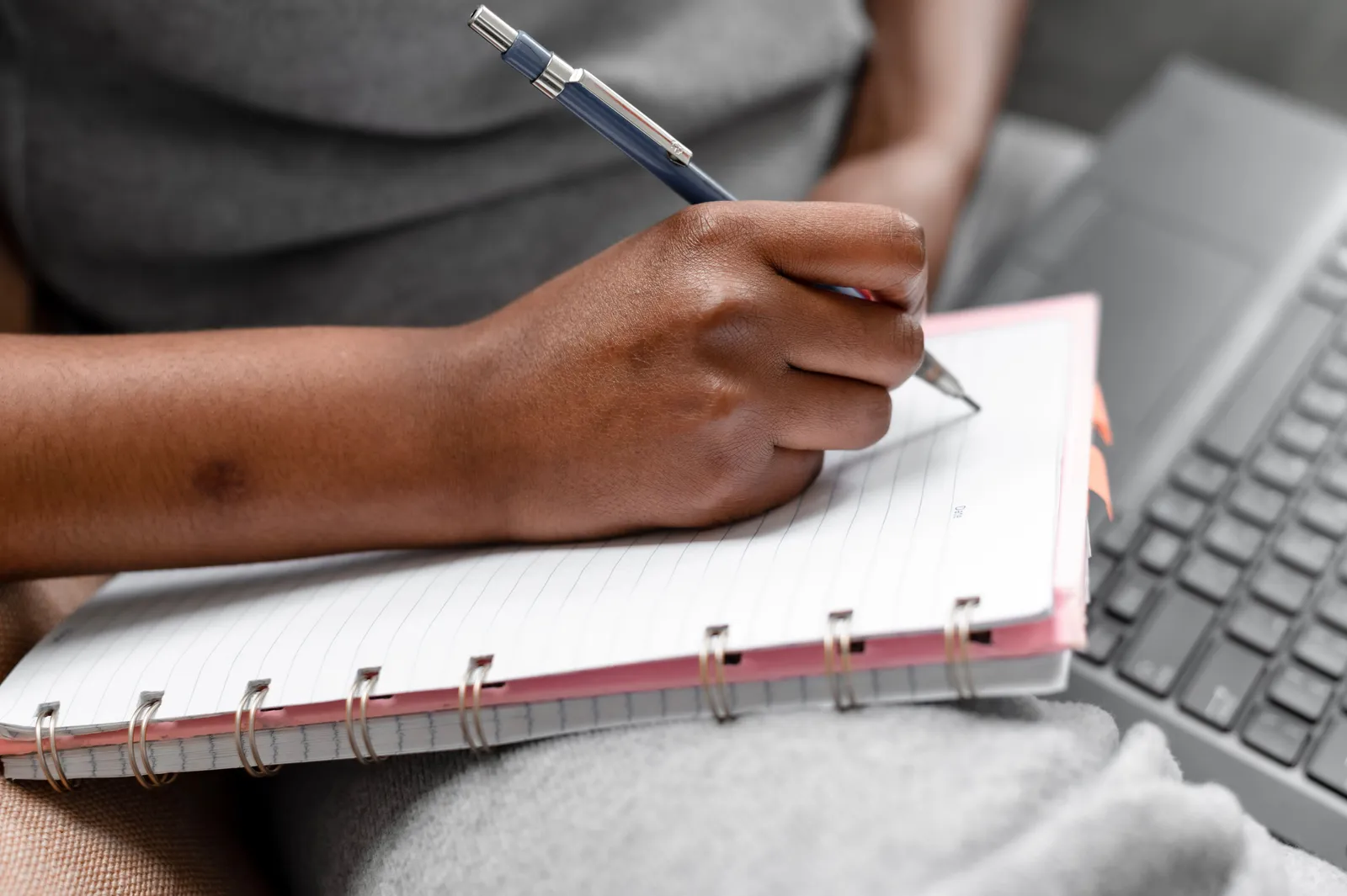 A person writing in a spiral notebook beside a laptop, focusing on note-taking and study