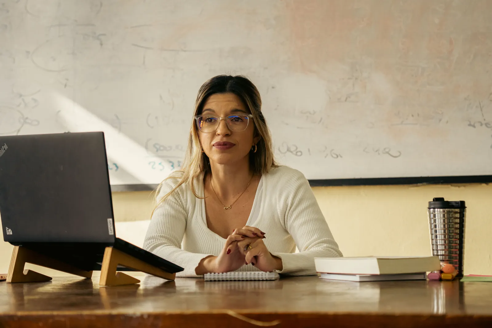 A woman teacher sits at a desk with a laptop and books in a classroom setting