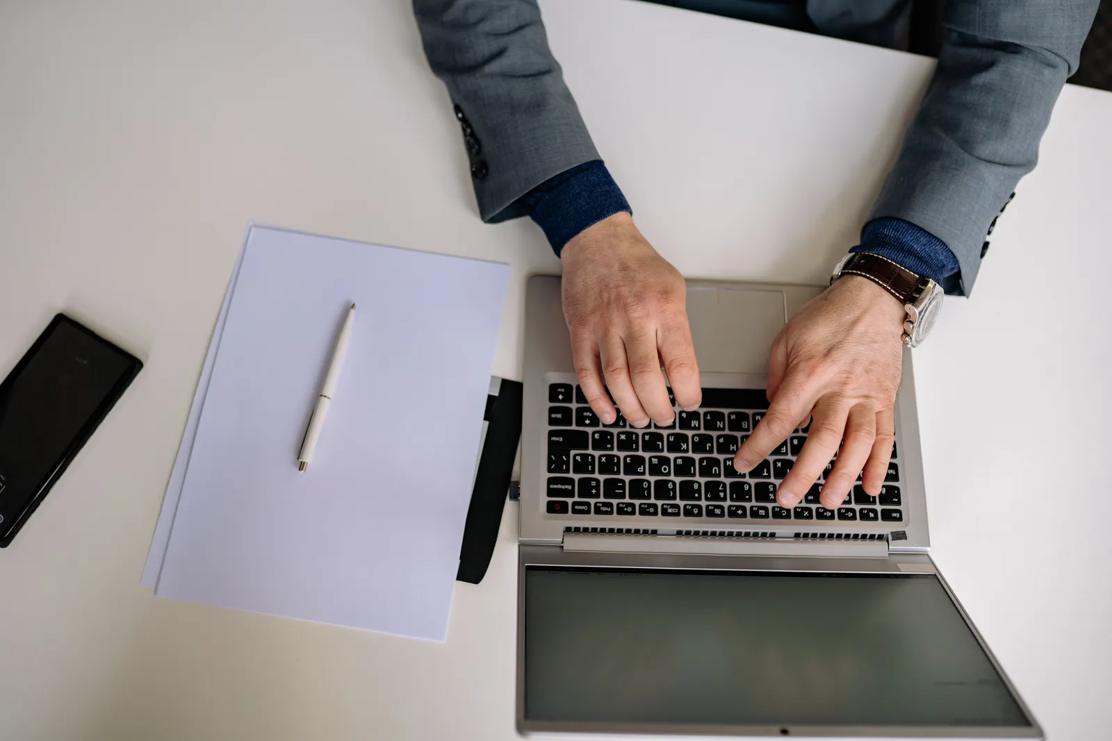 Overhead shot of a man in a suit typing on a laptop with a notepad and pen nearby