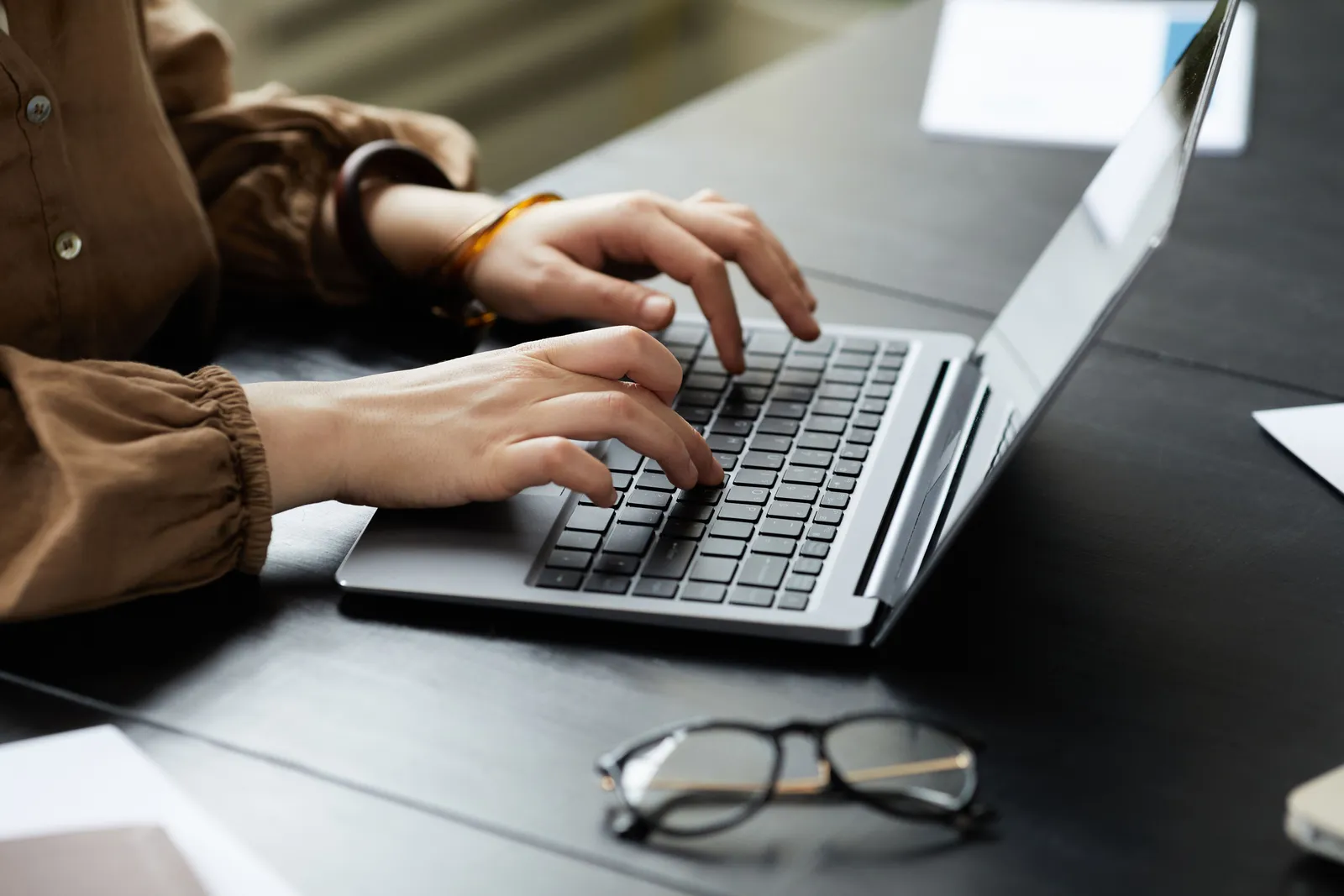 Hands typing on a laptop, eyeglasses on desk, modern workplace setting