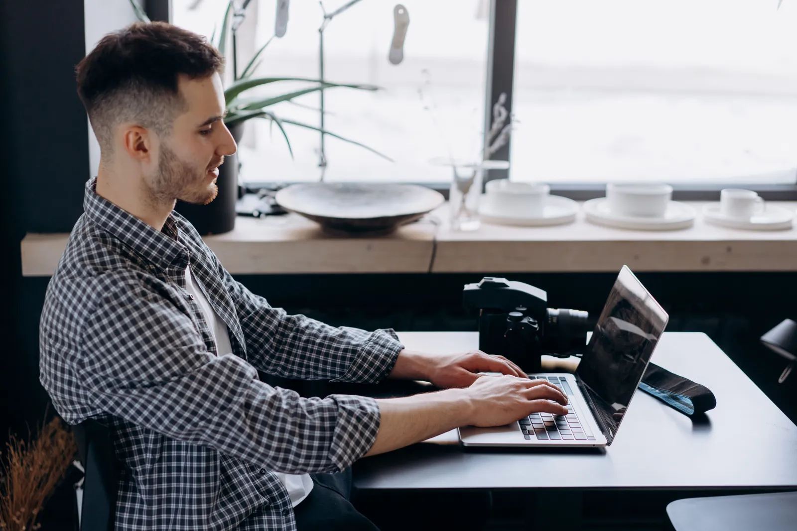 Young adult sitting at a desk with laptop and DSLR camera, working indoors