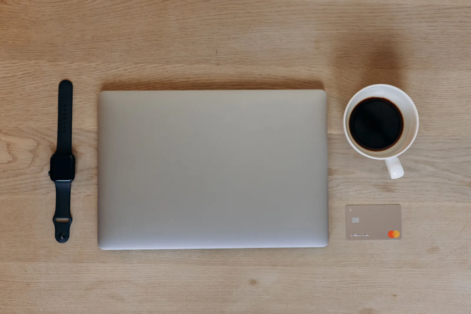 Overhead view of a sleek desk with a laptop, smartwatch, coffee, and credit card on a wooden surface