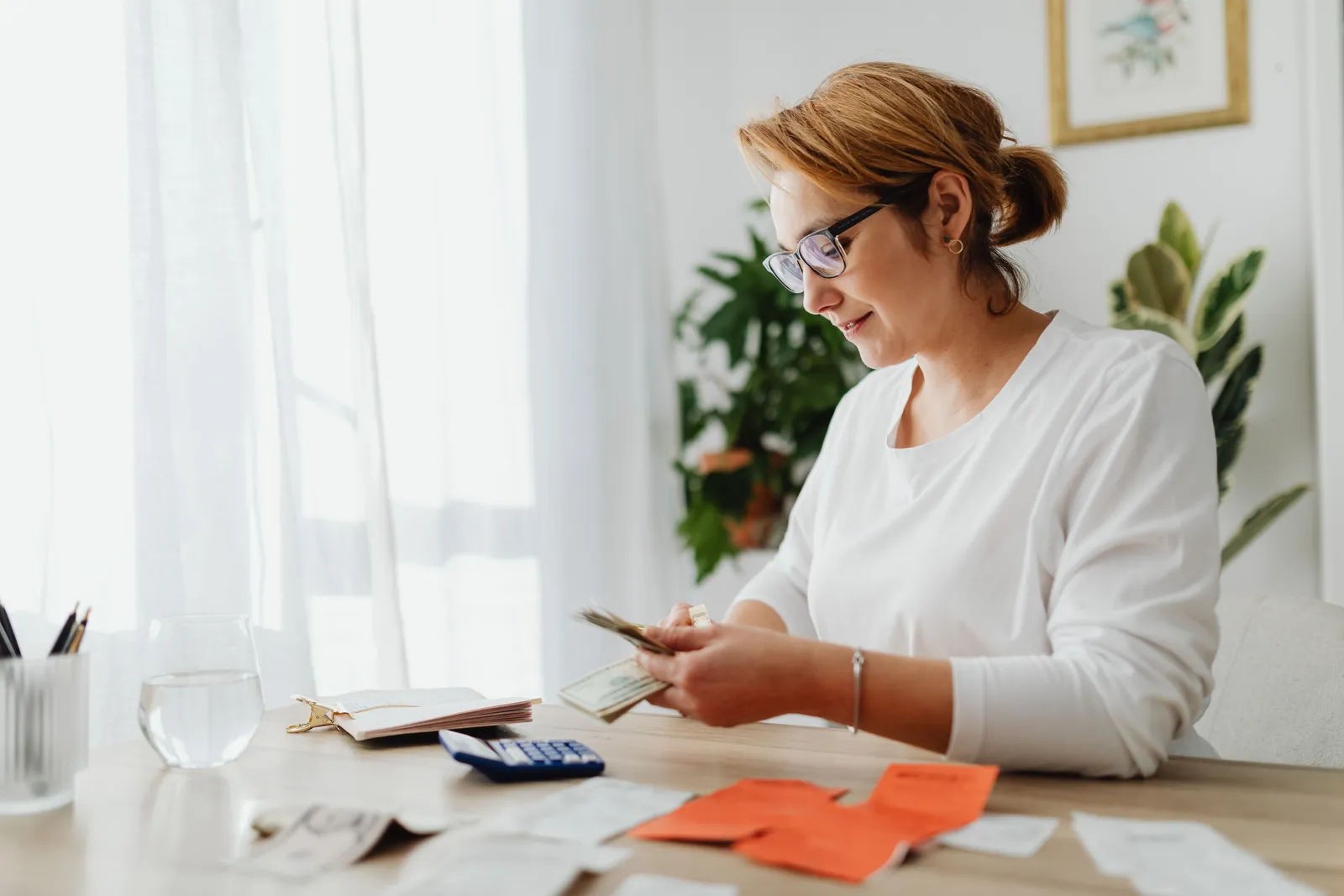 Woman counting money at home desk with papers and calculator, emphasizing financial management