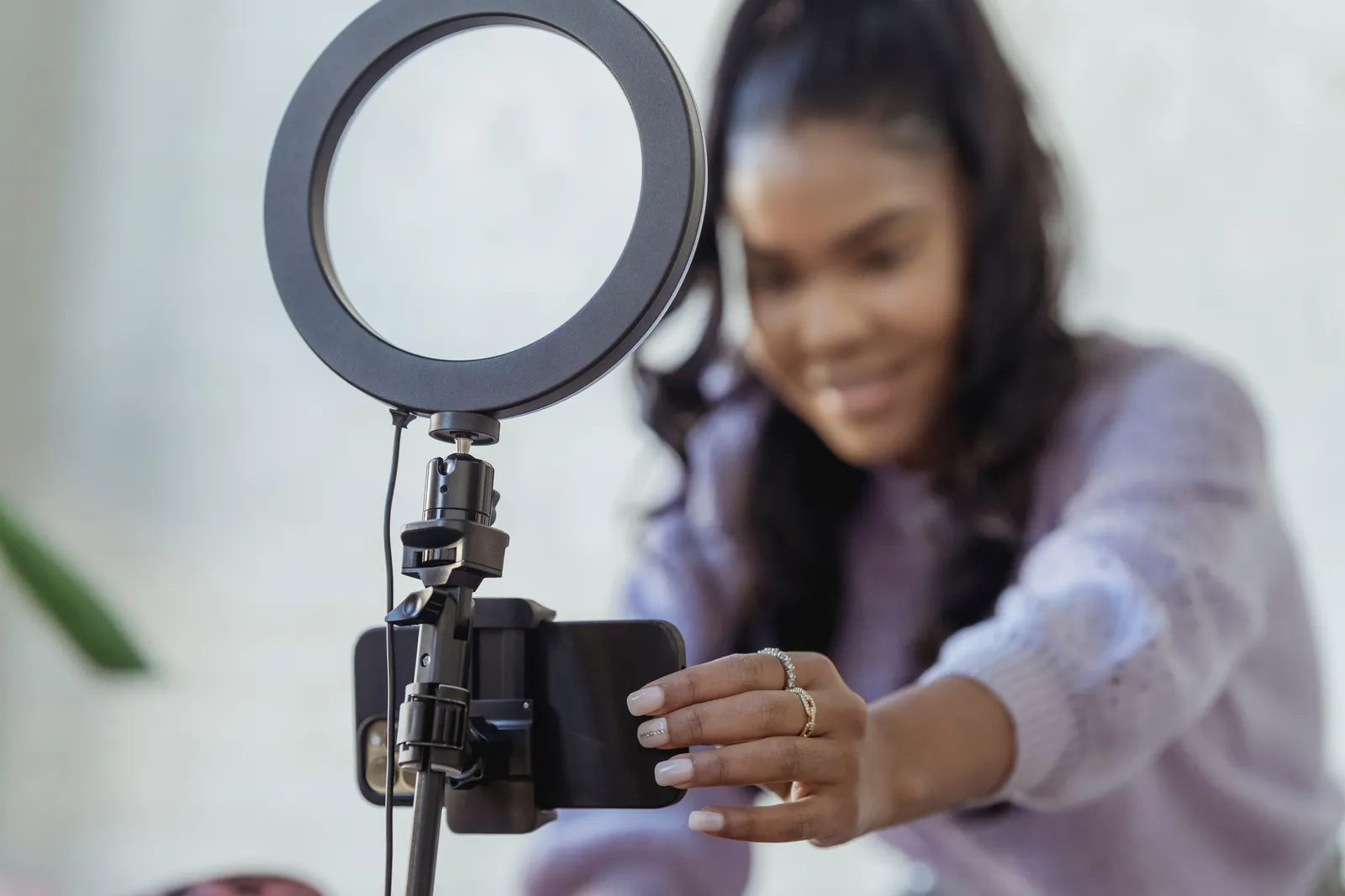 Cheerful young female blogger in stylish sweater smiling while setting up camera of smartphone attached to tripod with ring