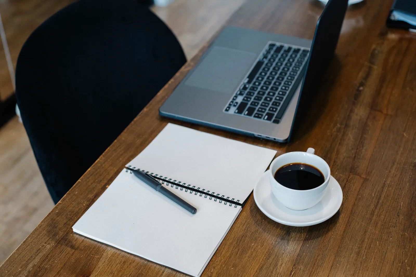 High angle of opened laptop and spiral notebook with blank pages placed on wooden table with cup of coffee and pen during