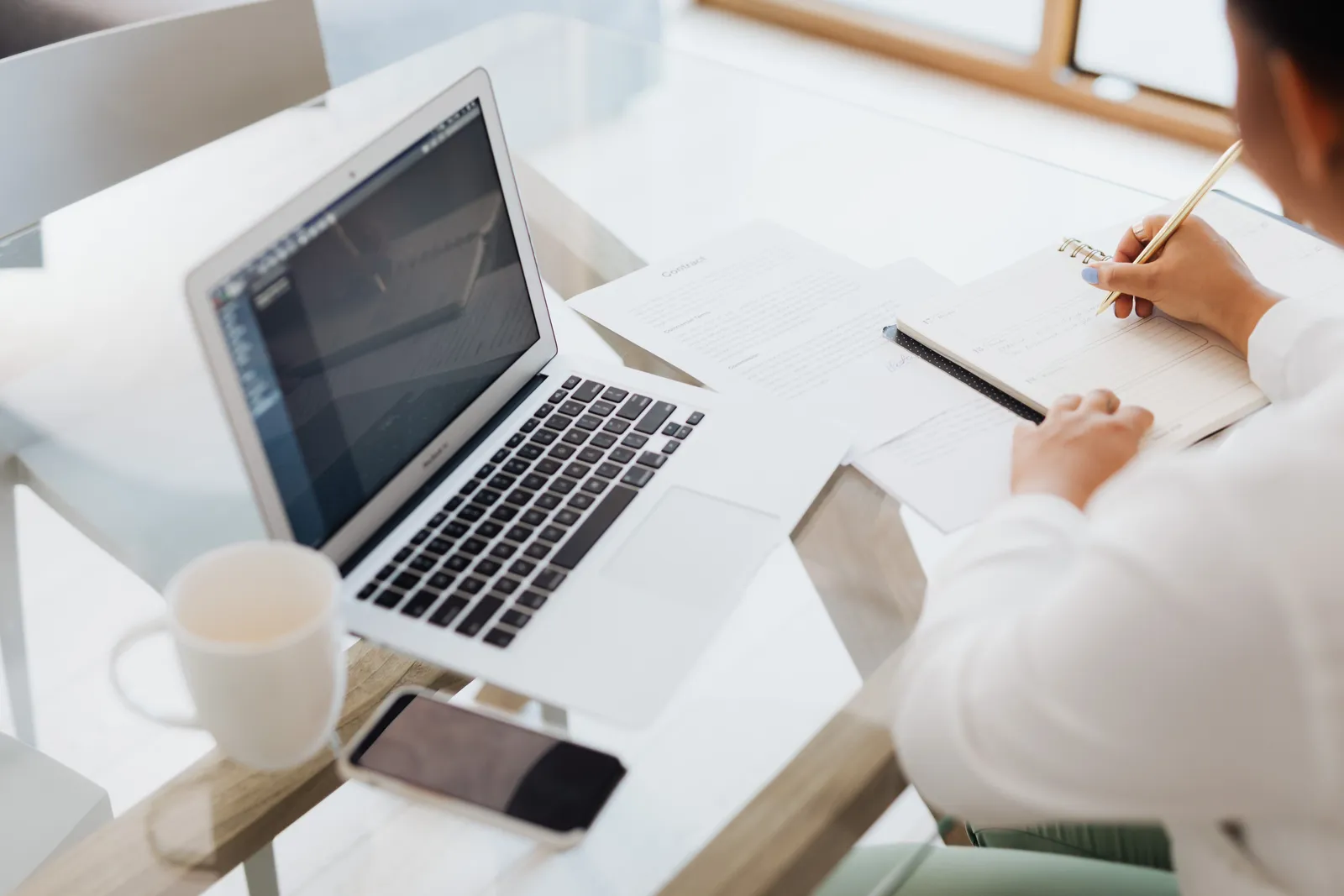 Person working at a glass table with a laptop, notebook, and coffee mug