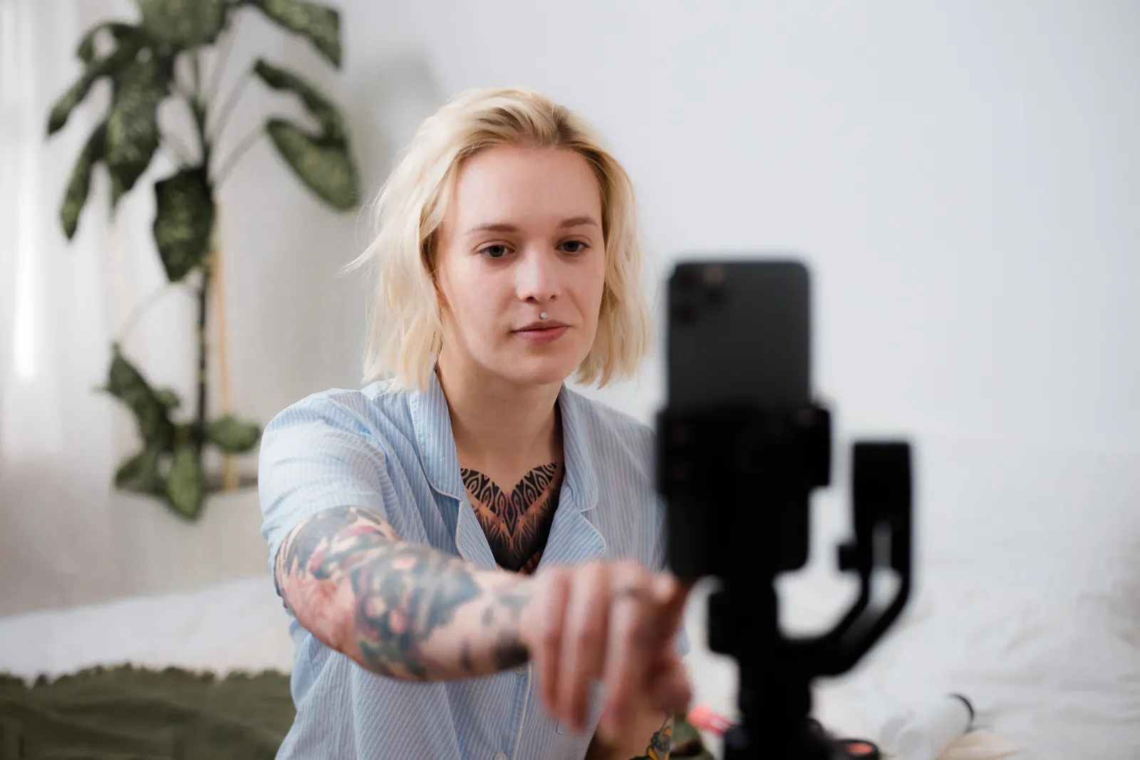 Tattooed woman with blonde hair adjusts smartphone on tripod for vlog setup indoors