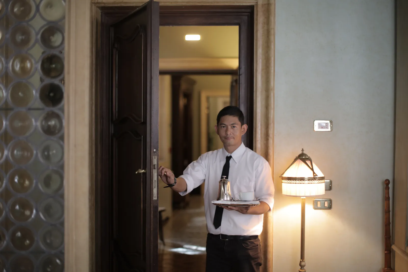 Friendly male waiter serving clients in room and carrying tray with coffee while working in stylish hotel
