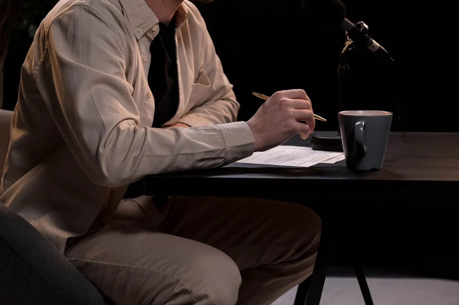 A man in a studio setting working on a podcast with microphone, pen, and coffee cup