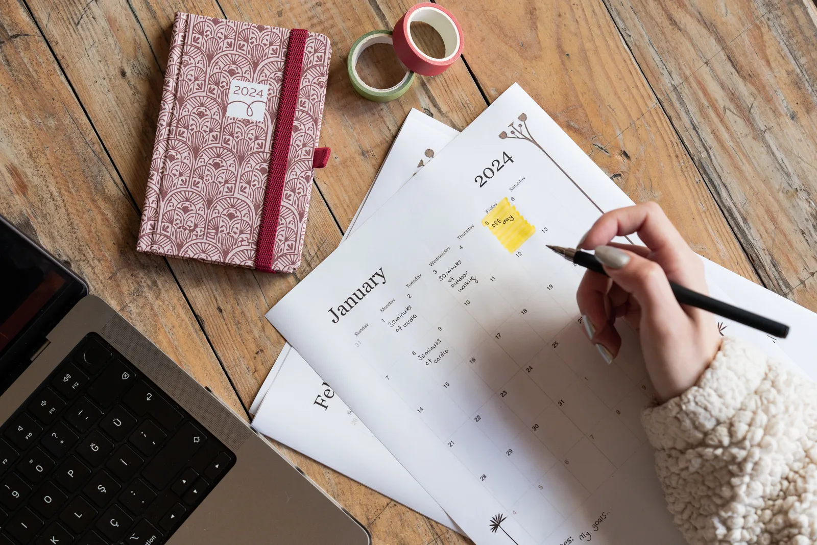 A person plans events in a 2024 calendar beside a notebook and laptop on a wooden desk