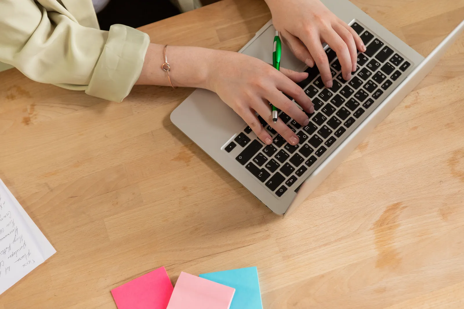 Hands typing on a laptop with a green pen and colorful sticky notes on a wooden desk