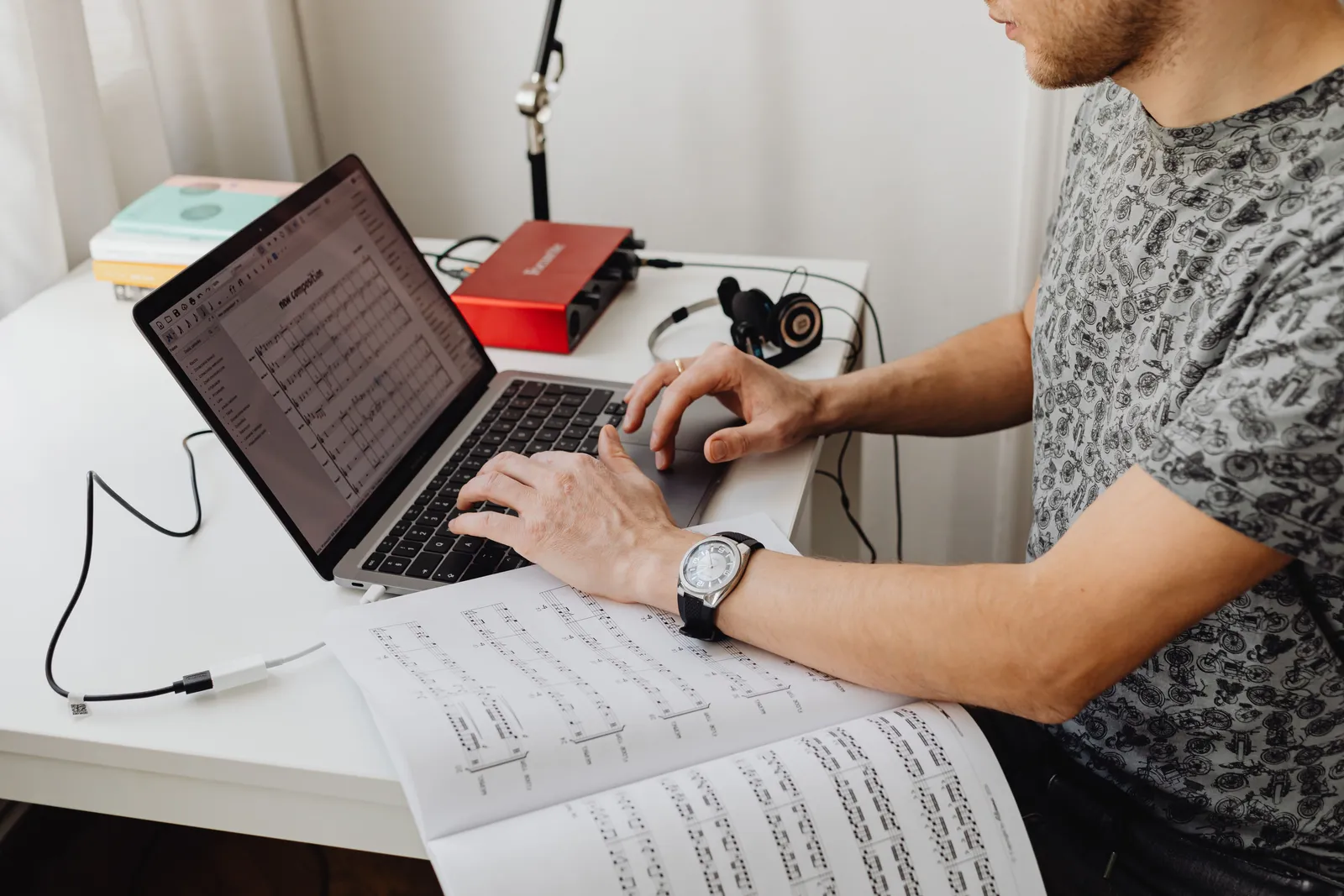 A man types music compositions on a laptop with music sheets spread across the desk