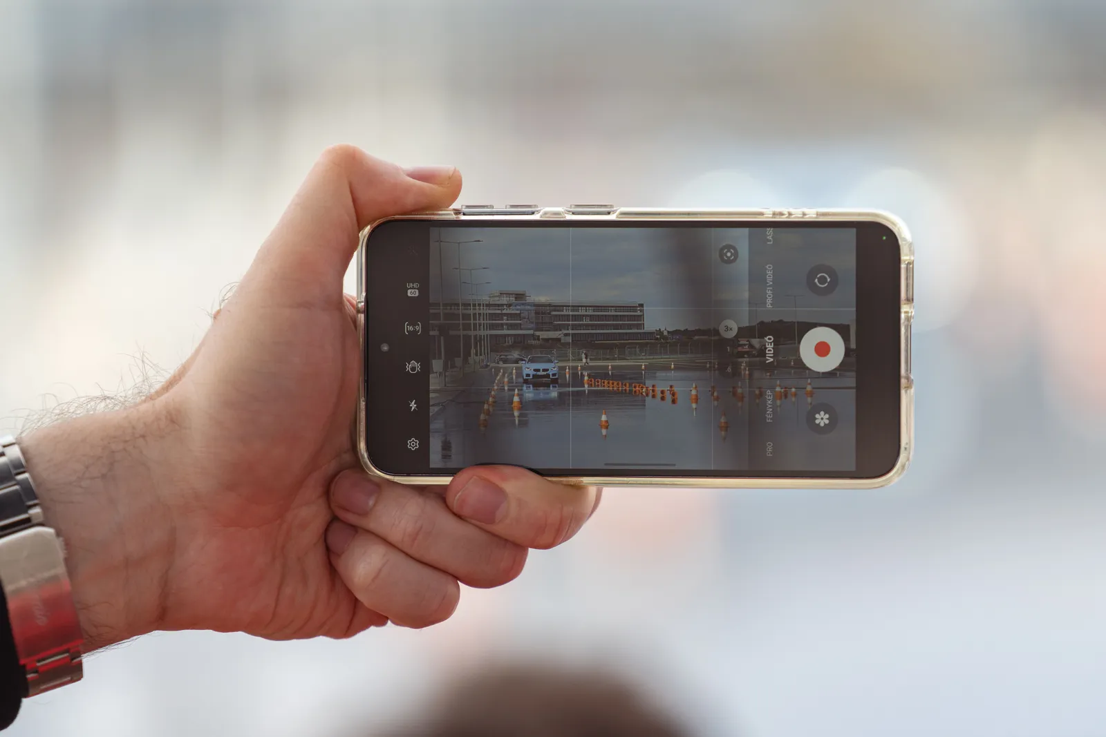 A hand holding a smartphone recording a video of a car scene outdoors. Captured in Balatonfőkajár, Hungary