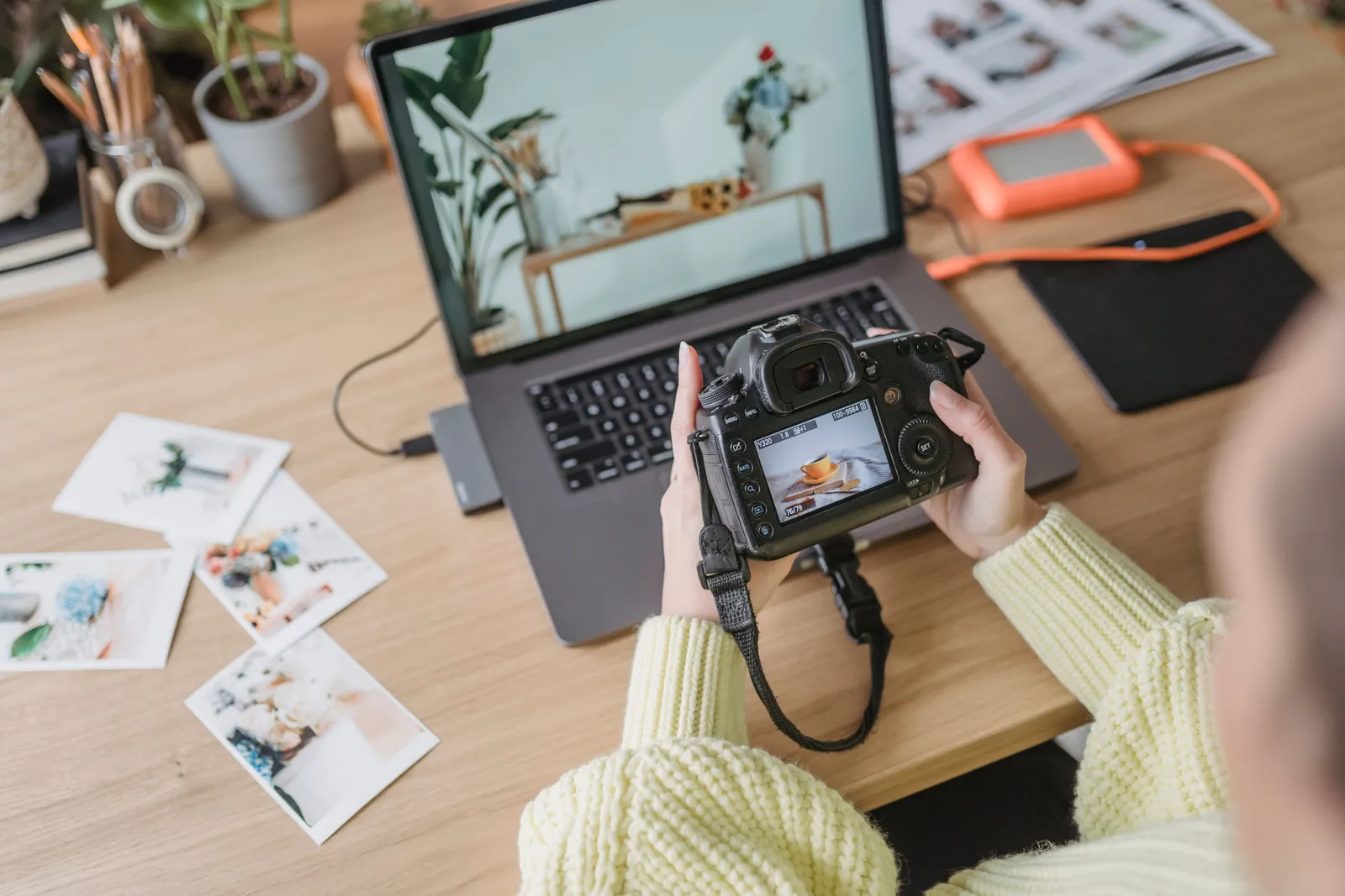 From above of crop anonymous female photographer with photo camera in hands sitting at table with laptop and printed photos