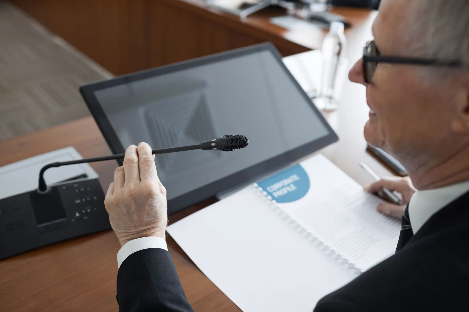 A senior businessman using a microphone during a conference meeting with documents and a monitor