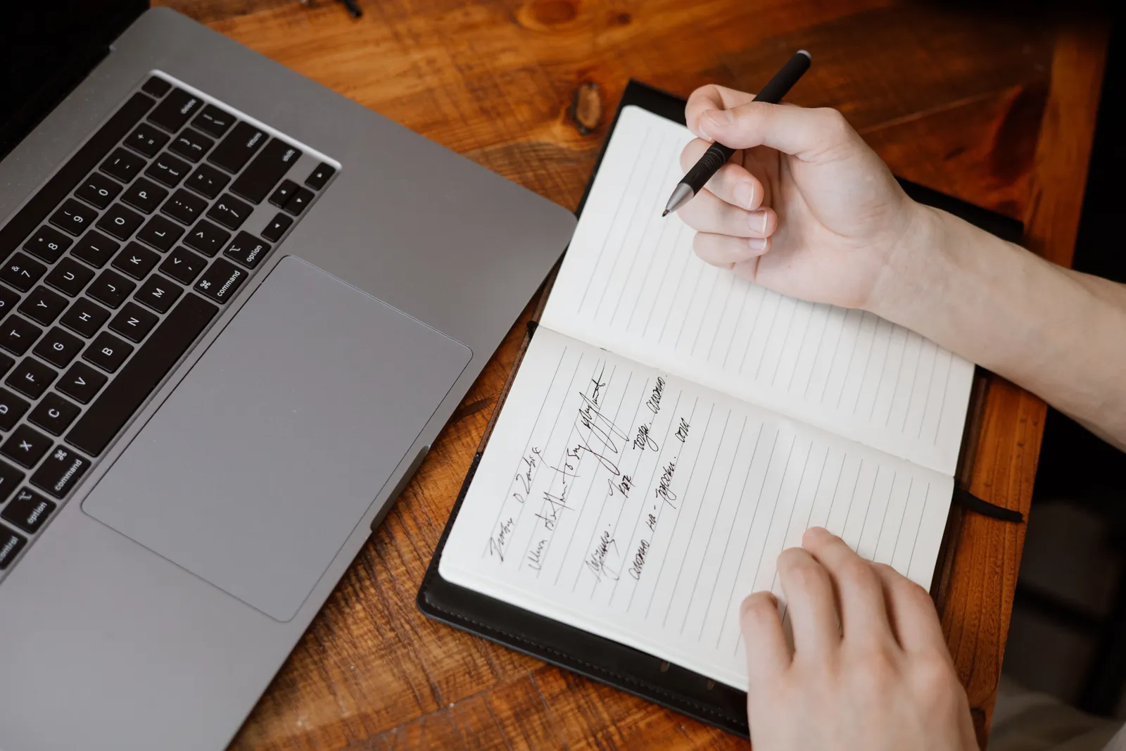 Close-up of hands writing in a notebook beside a laptop on a wooden table