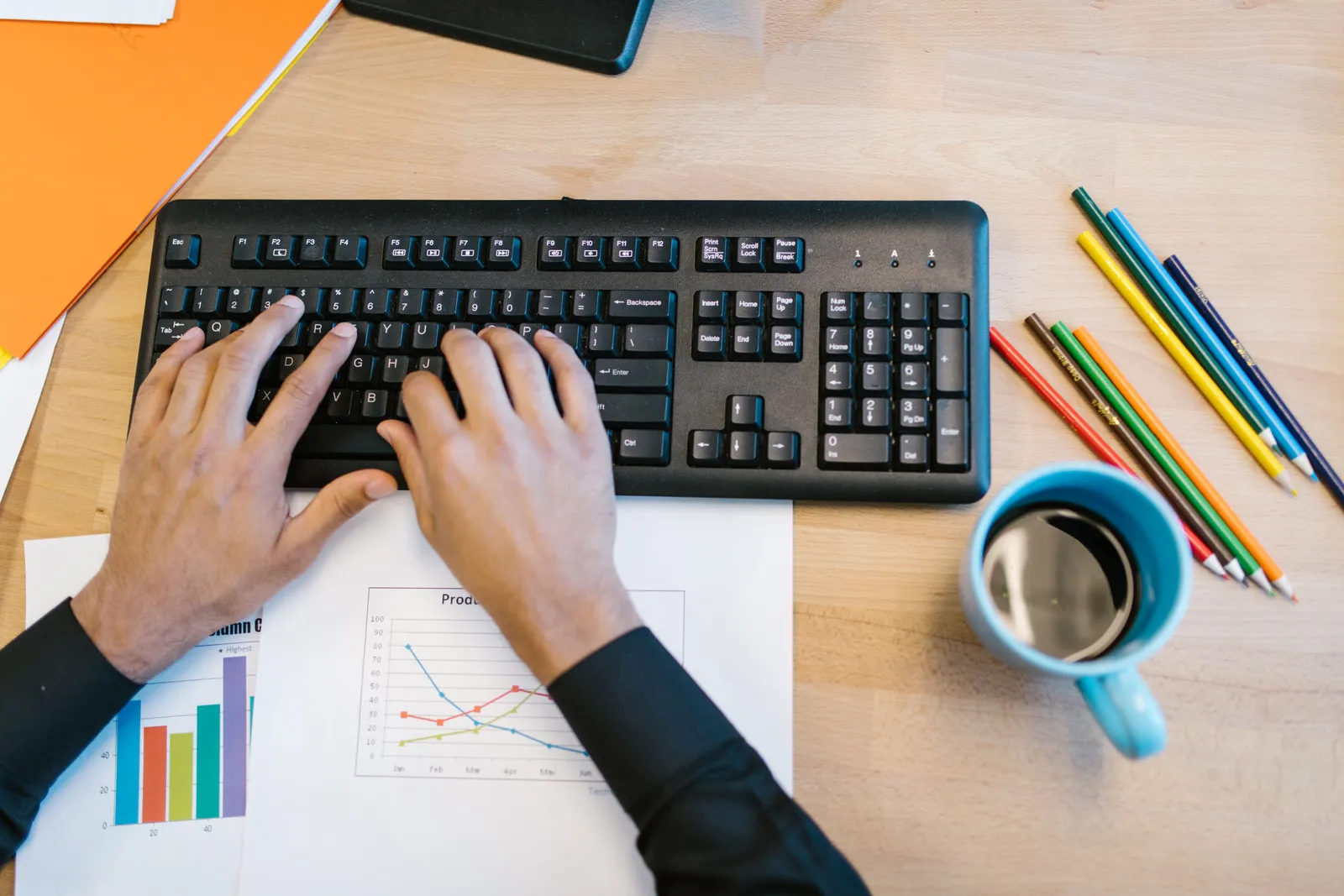 Workspace featuring hands typing on a keyboard, a coffee mug, colored pencils, and graphs on paper