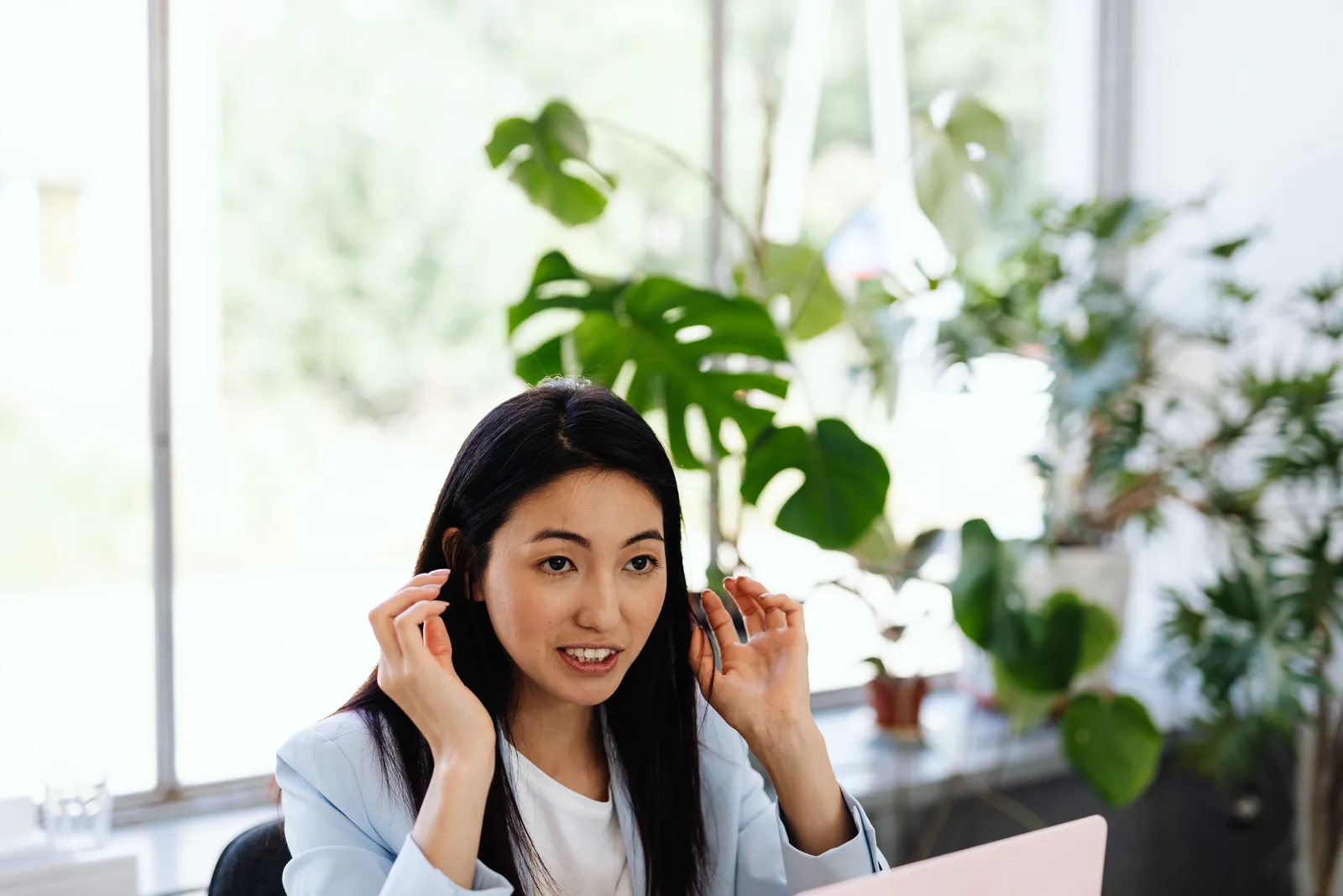 Woman in office, gesturing while speaking, surrounded by lush plants, near a window