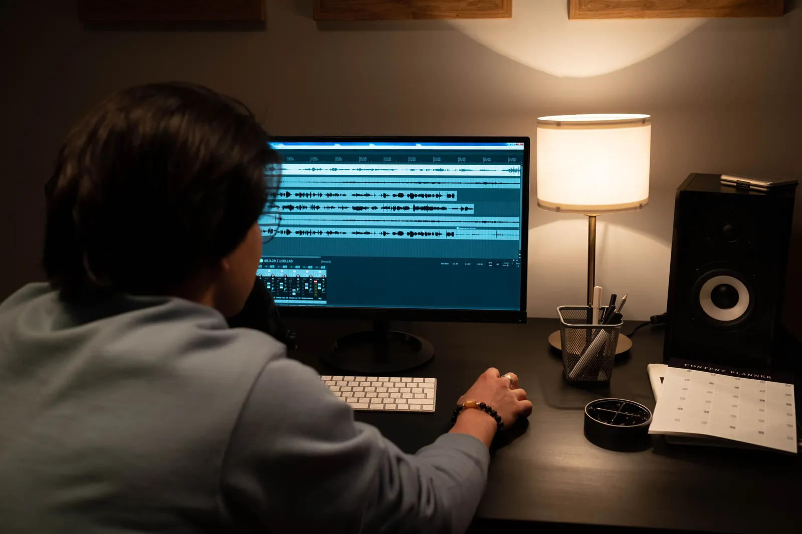 Man wearing a blue sweater editing audio on a monitor in a dimly lit room with a desk lamp