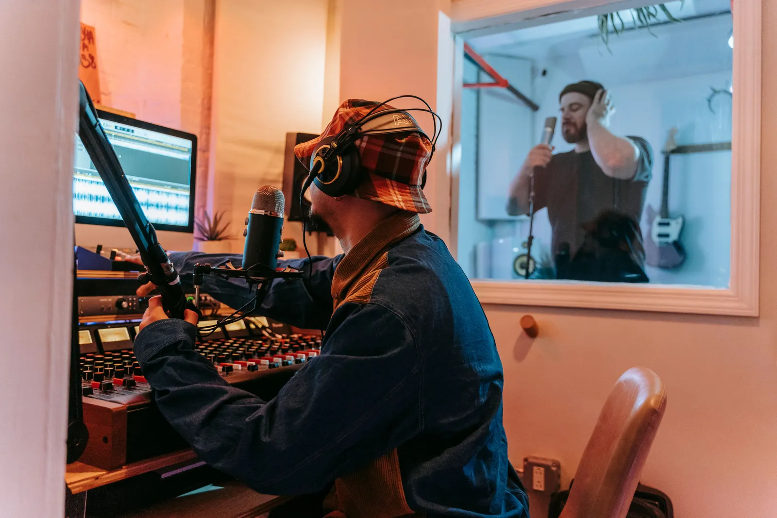 Man using sound mixer in studio with vocalist in soundproof booth for recording session