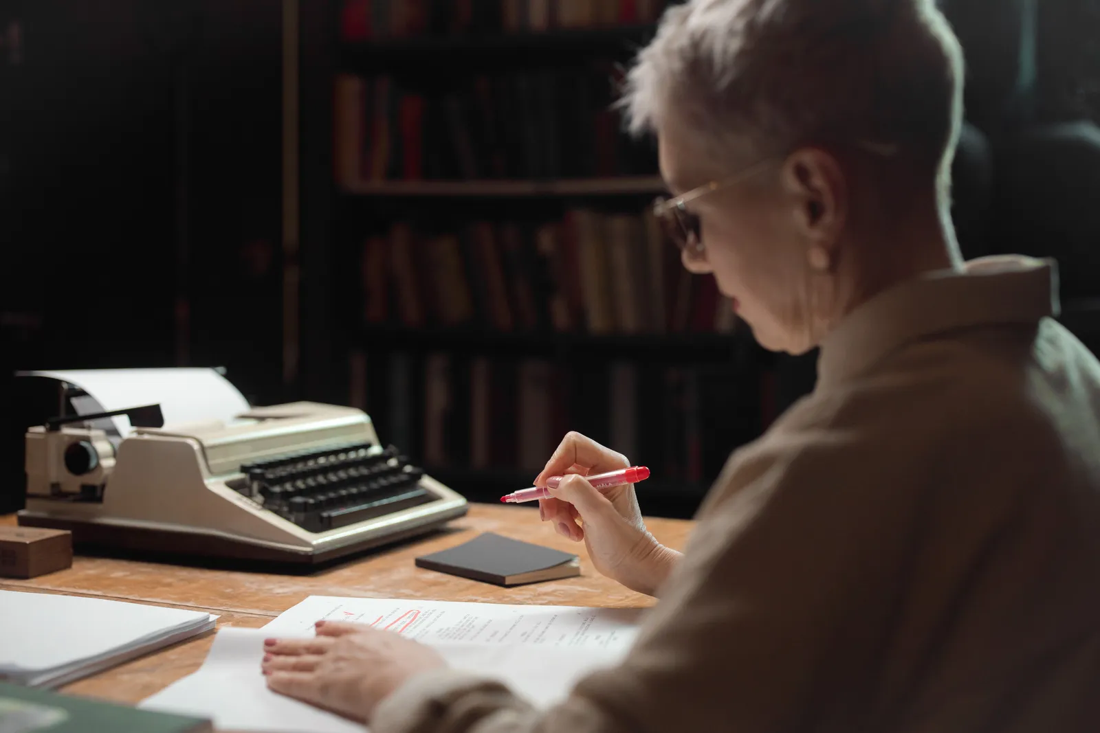 Elderly woman reviewing a manuscript with a typewriter at a wooden desk