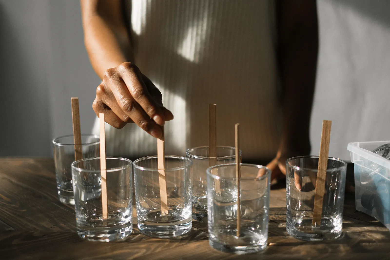 Unrecognizable female securing wooden wicks in glass molds while making aroma candles at wooden table in light studio