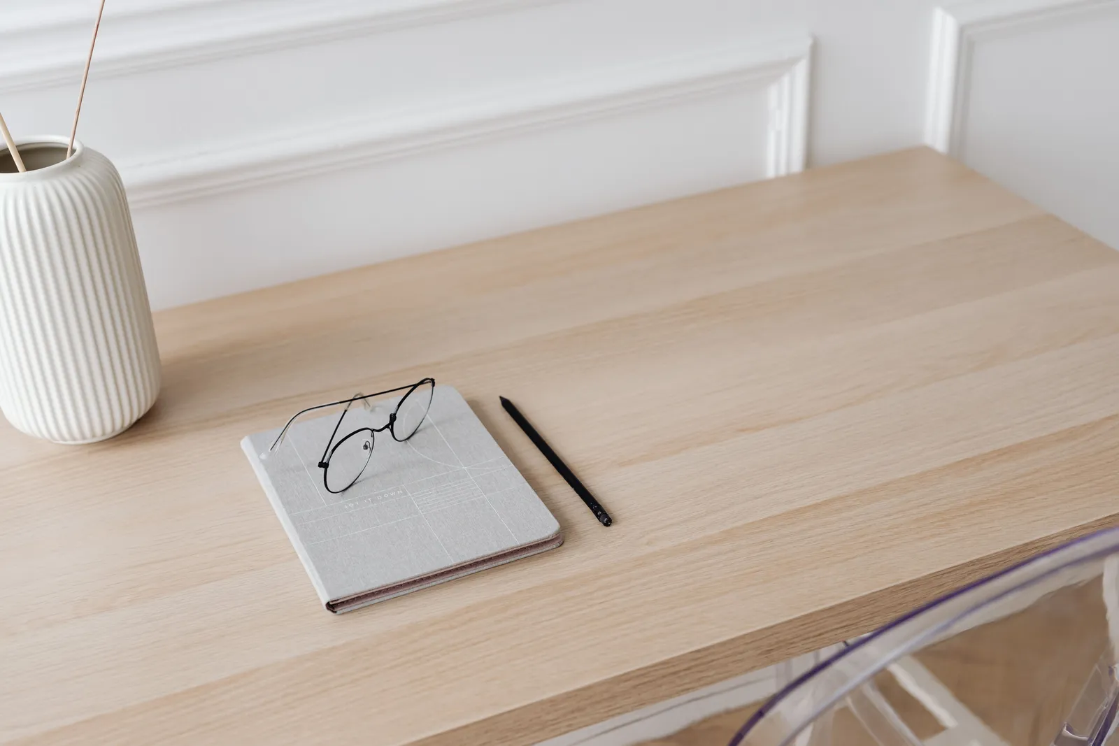 A minimalist workspace featuring a notebook, eyeglasses, pen, and a decorative vase on a wooden desk